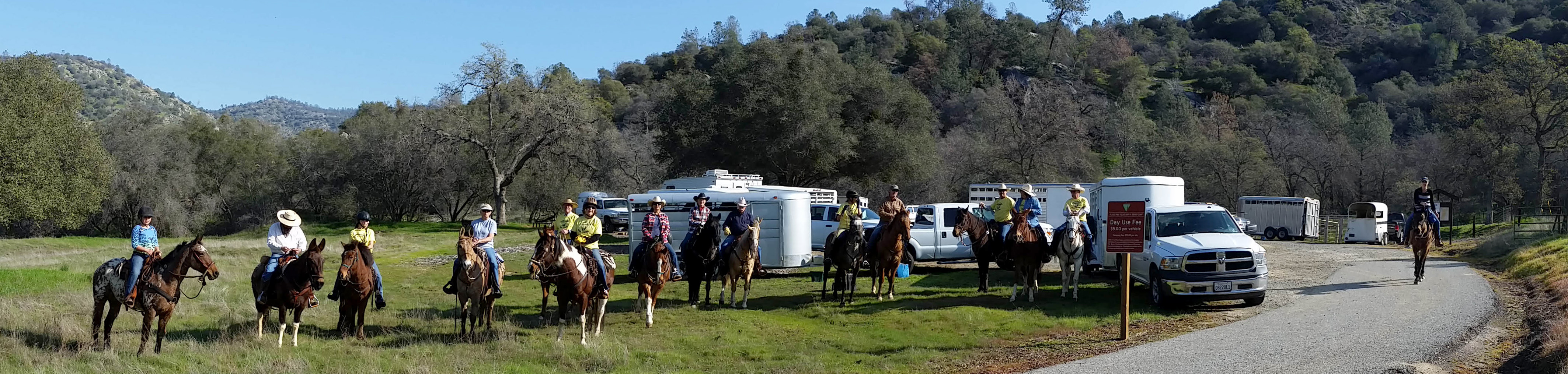 Riders on horses pose in front of a parking area and oak covered hillsides.