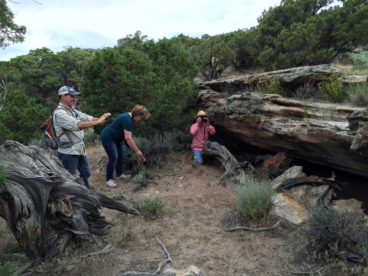 Three people are next to a large rock with a cave-like opening underneath. Two of them have cameras, taking photos.