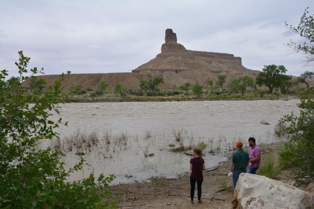 Three individuals stand in front of a large river with rocks, trees, and the sky shown.