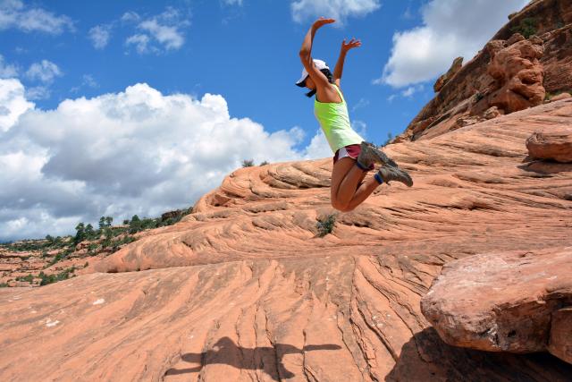 Girl jumps in the air on a rocky surface with blue sky and a few clouds in the background