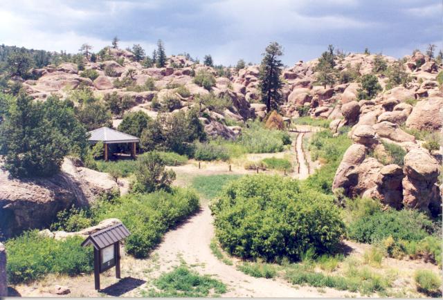 A kiosk, shelter, and trail with cable fencing are visible among rounded granite boulders in Penitente Canyon.