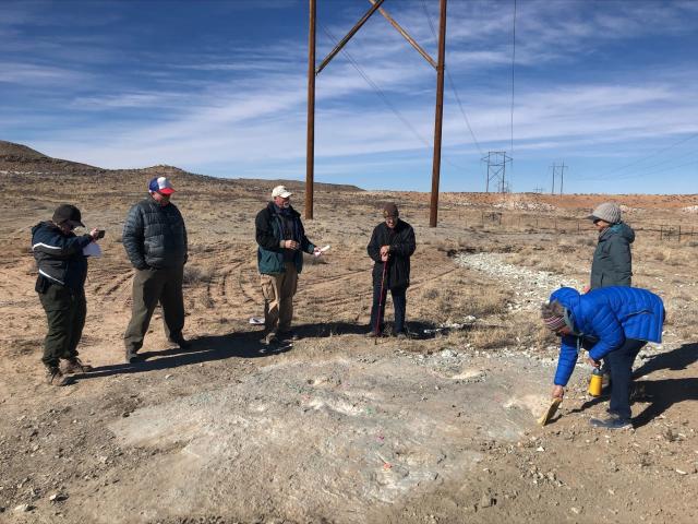 Five individuals standing and one bending over brushing the tracksite. A telephone pole is in the background. The ground is dry, flat, and has little vegetation.