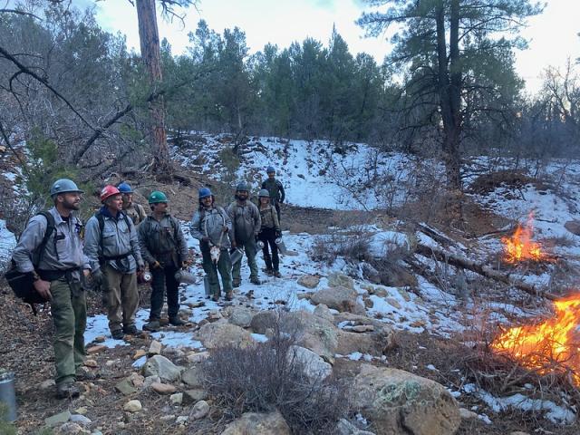 A line of people in protective gear stand in the snow during a prescribed burn.