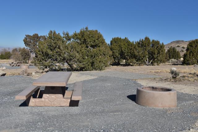 Campsite with a picnic table, fire pit, and tent pad. The ground is gravel and the background has juniper trees and a blue sky.