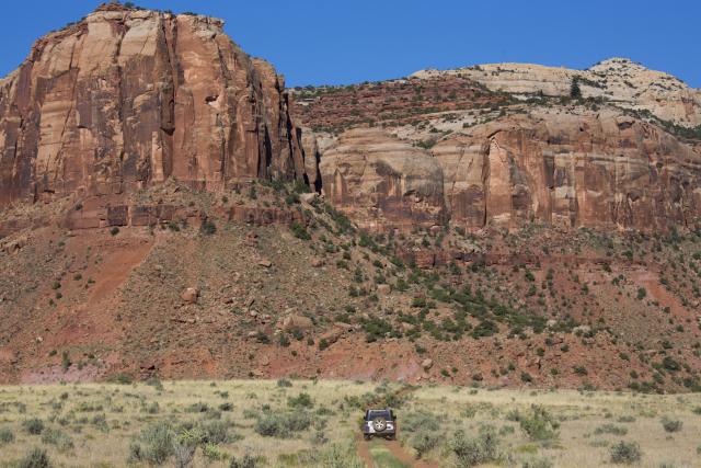 A vehicle drives down a two-track road with red sandstone cliffs looming.