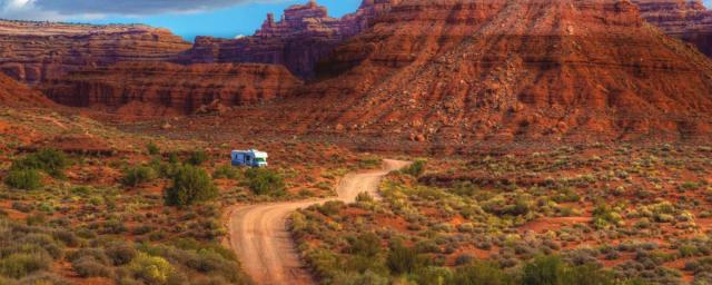 An RV driving on a dirt road through the Monticello -  San Juan River Area and red rock formations in the background. 