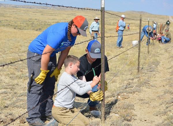 A volunteer looks on as two younger volunteers work with a tool on the fence. 