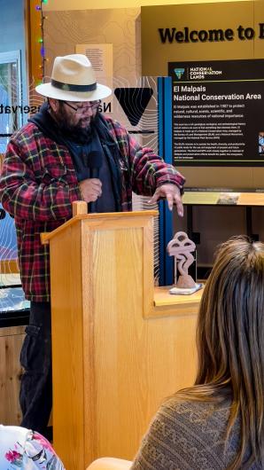 Walter Torres speaking with a microphone at a podium. He is pointing at a scaled down version of his sculpture which depicts a volcanic eruption. A woman listens. 