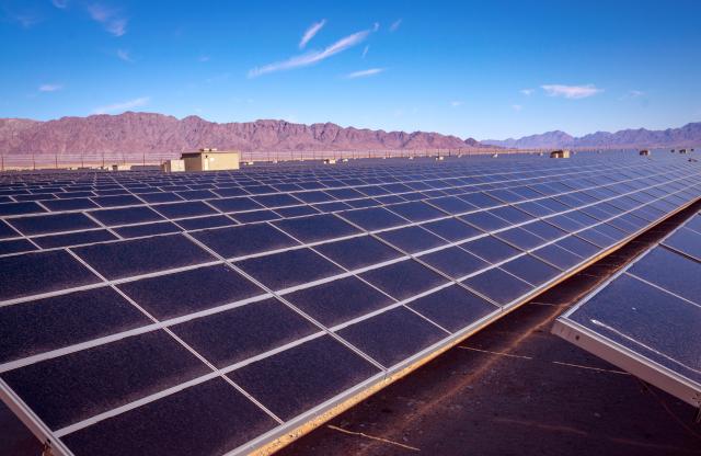 A long row of solar panels in an open area, with a mountain range in the background.