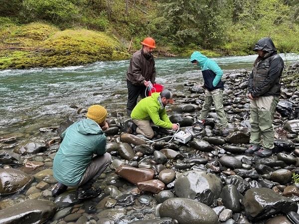 5 people standing or kneeling in large stone next to a stream. One of them is handling a device. 