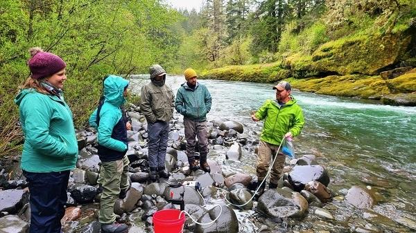 5 people standing in large boulders next to a stream. A man is holding a cord that connects to a white device. 