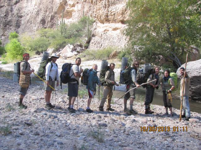 Nine people standing on the bank of a creek with some of them carrying unique fish traps on their backs