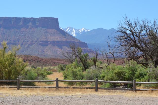 Gravel road, wood fence, trees, rocky mountain and blue sky.