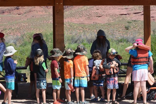 Youth lineup near covered picnic tables for orange lifejackets.