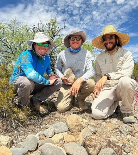 Three men smiling behind stones organized into a mound. The man in the center is holding a stone. 