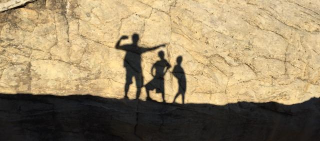 The shadows of three children dancing appear on a yellow sandstone wall.