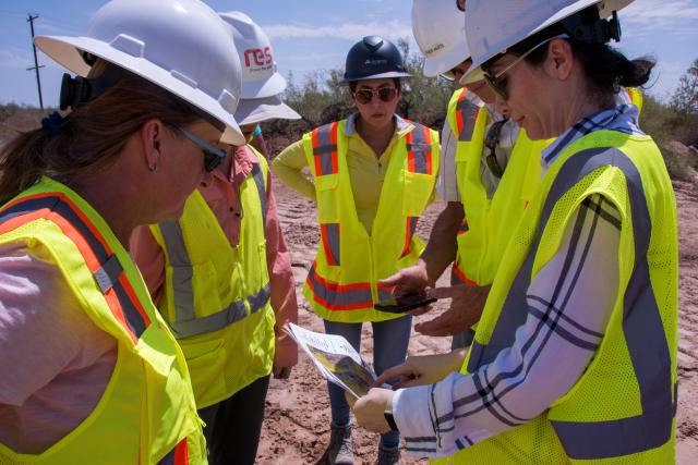 A group of people dressed in hard hats and bright yellow safety vests huddle around a phone and a map.