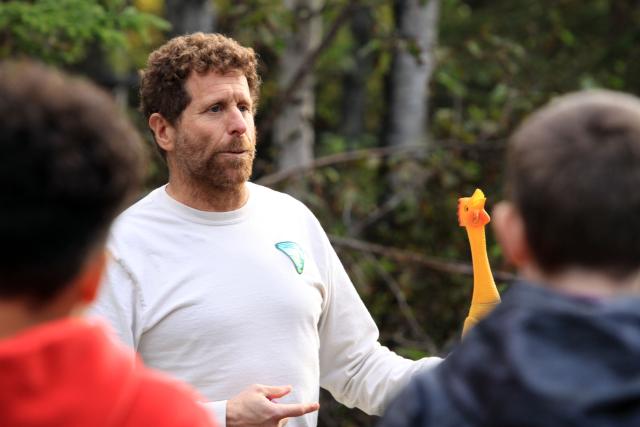 A man wearing a beige t-shirt stands outside holding a rubber chicken while the man speaks to young students.