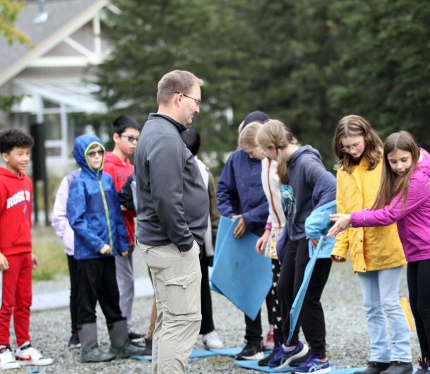 A man watches a group of children pass blue foam mats from one person to another.