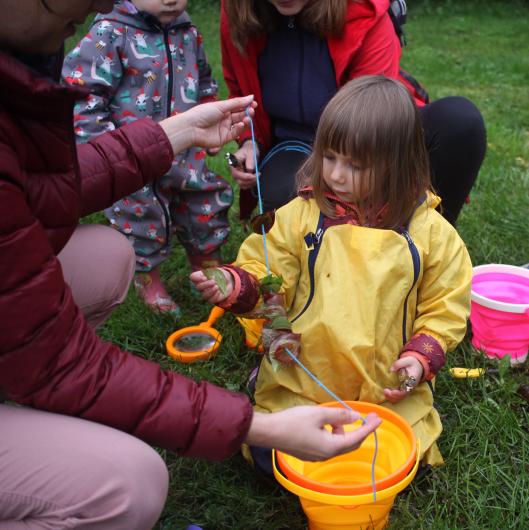 A small girl wearing a bright yellow rain coat helps her mom string leaves on a blue cord.