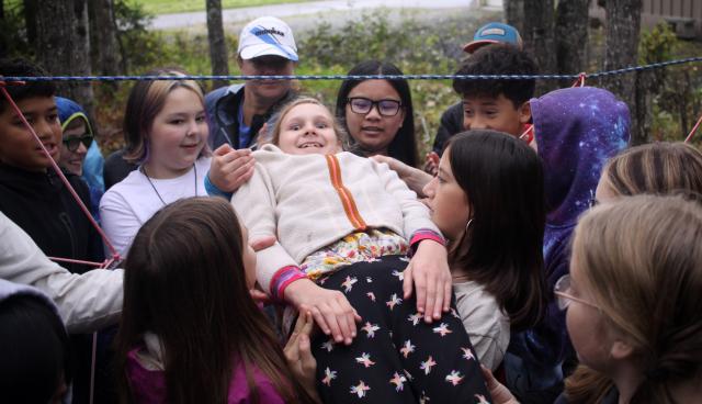 A group of students stand outside and help pass a classmate up and through a hole in a make believe spider web. The web is made out of rope strung between two birch trees.