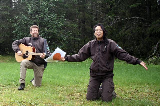 A man is on bended knee in the green grass playing a guitar while a woman with outstretched arms kneels in the same green grass singing