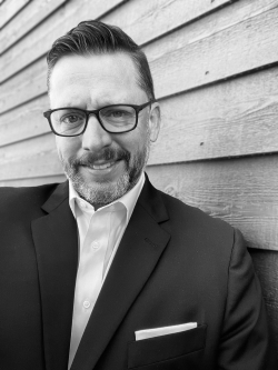 A black and white photo of John Gale with short dark hair and glasses wearing a suit jacket leaning against a wall