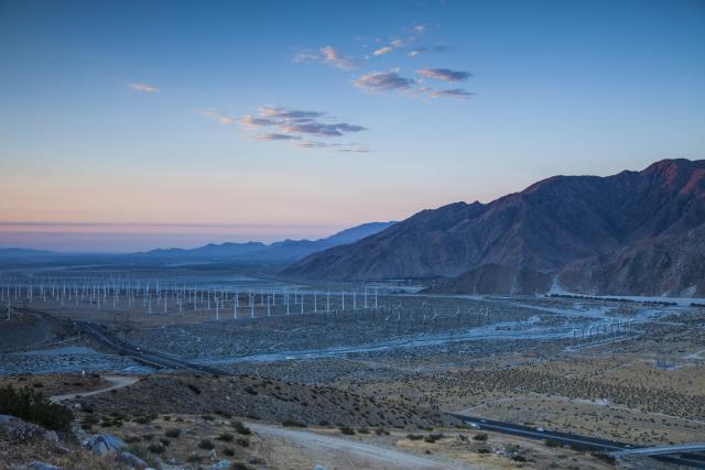 Wind turbines lined across a landscape with scattered vegetation and mountains in the background. 