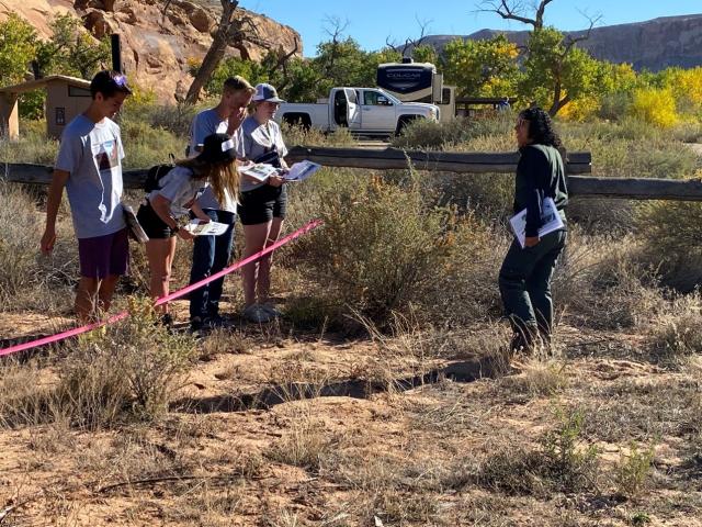 Student stand in a group learning about wildlife out on the landscape with a Forest Service Biologist.