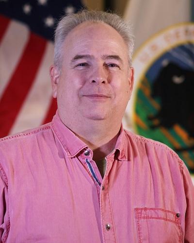 person with short hair wearing a collared shirt posing in front of the American flag