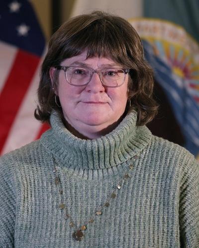 person with dark hair wearing a turtleneck sweater and necklace posing in front of the American flag