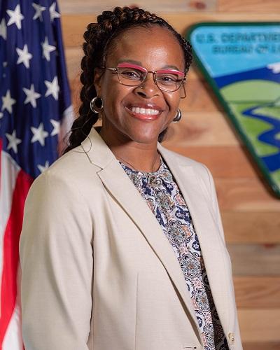 person with dark hair and glasses wearing a printed shirt posing in front of the American flag