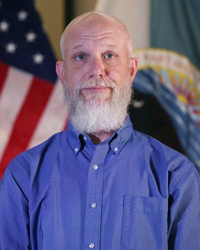 person with short hair and beard wearing a collared shirt posing in front of the American flag