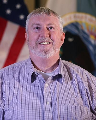 person with short hair wearing a collared shirt posing in front of the American flag