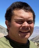 person with short dark hair and green shirt posing in a mountainous landscape