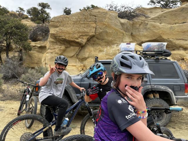 A photo of a group of youth mountain bike riders sitting atop their bikes on a desert trail, with an evening cloudy sky in the background.