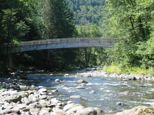 Photo shows a bridge crossing the Wild and Scenic Salmon River in a mountainous, forested environment with river rocks in the foreground.