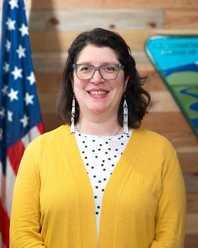 smiling person with glasses, polka dot shirt, and a yellow sweater posing in front of the american flag and blm logo