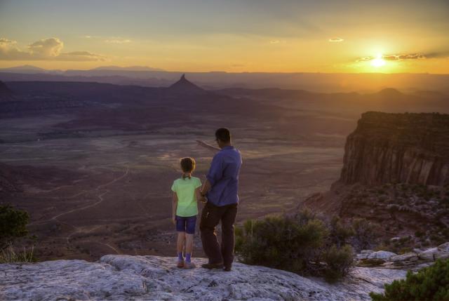 Photo shows a man with a blue shirt and brown pants holding a child’s hand, while standing on the edge of a canyon overlooking a desert valley landscape. The child is wearing a lime green shirt and blue shorts. The sun is setting in the background. 