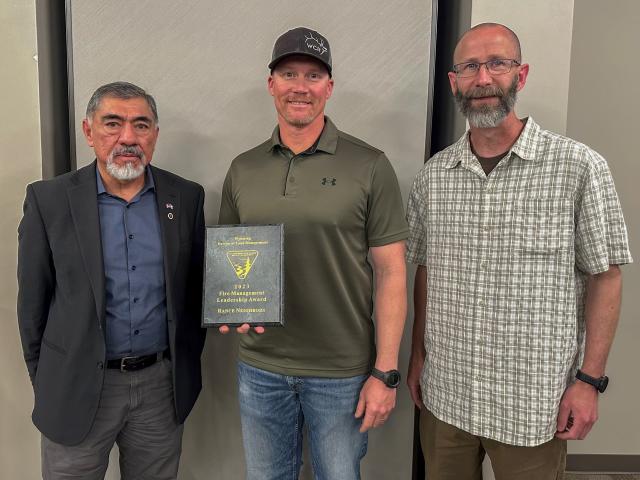 Three people stand smiling at the camera. The person in the middle is holding a plaque.