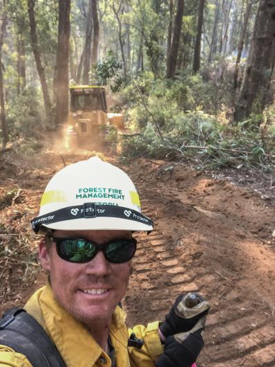 A person wearing sunglasses and hardhat takes a selfie in a forest with an excavator digging fireline behind him.