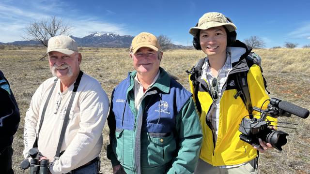 Alt text: The photo shows three men facing the camera in outdoor gear as they stand in a grassland area with mountains in the background. Liem is on the right, wearing headphones and holding a video camera.  