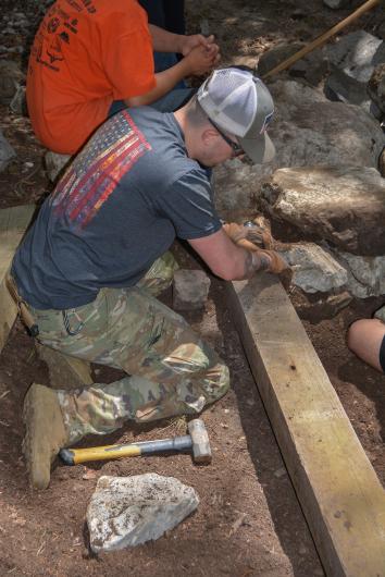 A person wearing gloves, sunglasses and a ballcap crouches on the ground as he works to place a wooden post, or ballast, along the perimeter of a campsite.