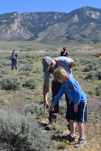 A child and an adult point to something they see on the ground in an open landscape with scenic mountains in the background.