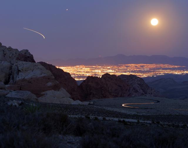A bright moon shines above layered rock formations and the glowing lights of urban developments at twilight.