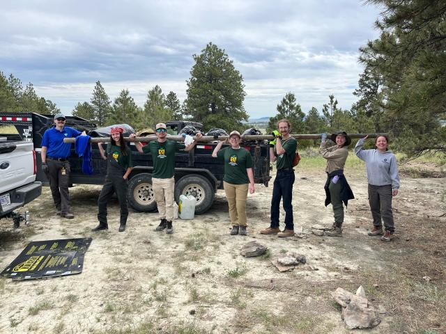 Seven people in a row holding a long pole on their shoulders. Trailer full of trash behind them. 