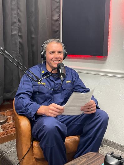 A Caucasian adult man in a blue jumpsuit sits in headphones and a microphone smiling at the camera.