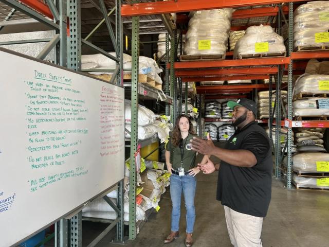 African American man reads aloud from a white board titled "Daily Safety Items" to a listening Caucasian woman. 