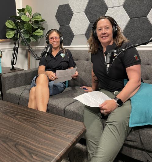 Two adult Caucasian women sit on a couch with microphones and headphones