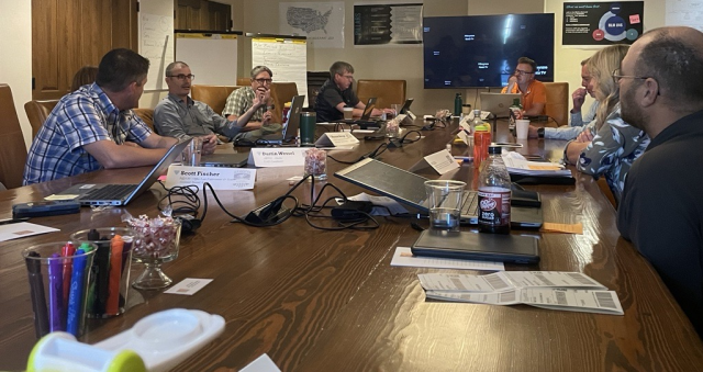 Caucasian women and men gather around a table with laptops and papers on it.